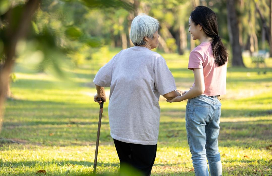 An older woman with a cane and a younger woman walking together in a park. 