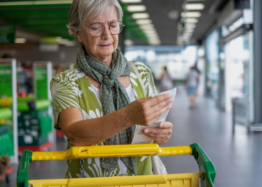 A senior woman outside the supermarket checks her grocery receipt looking worried.