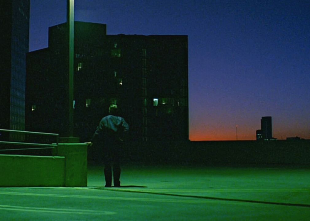 A solitary figure stands in a parking lot under a green light with a sunset in the distance