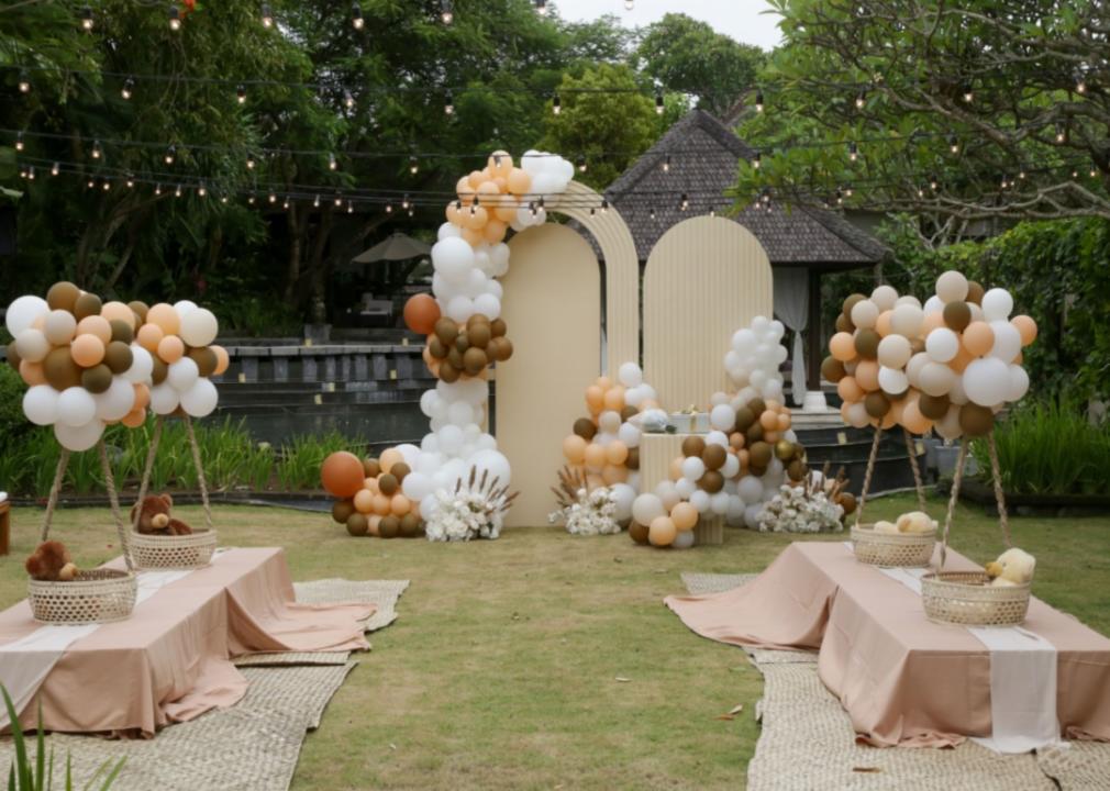 A baby shower set up outdoors with string lights, baby baskets with stuffed animals atop of low tables, blanketed seating on the floor, and white, brown, and tan balloons tied in bunches.