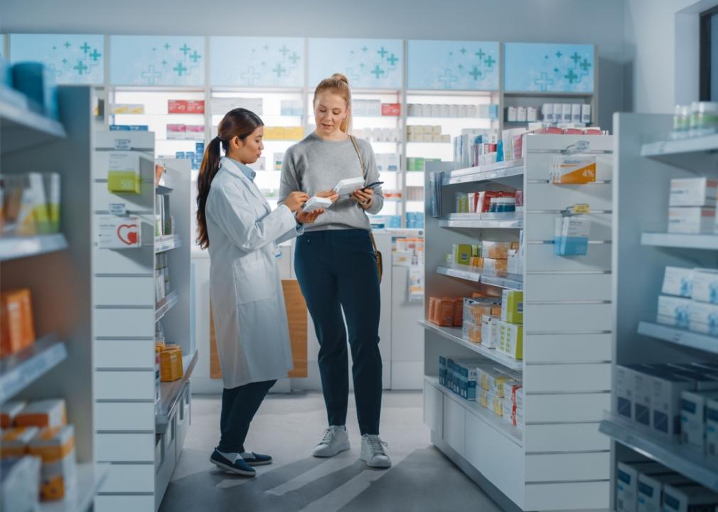 A statuesque caucasian woman stands alongside a pharmacist in a pharmacy holding a pack of medication. 
