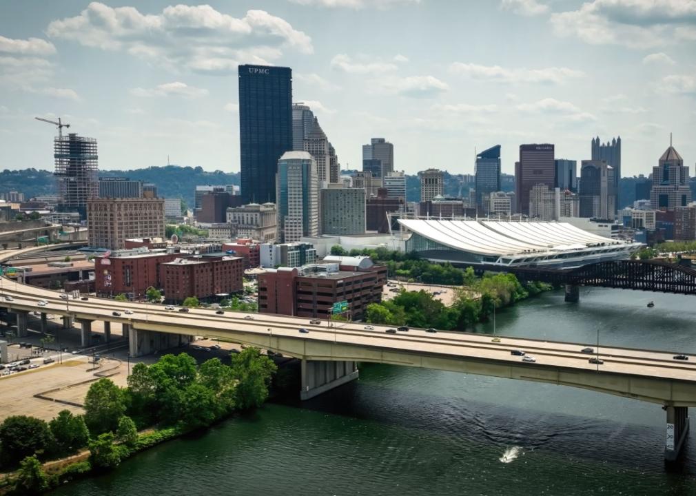 Aerial view of a city with a river flowing through it. The city has a mix of modern and historic buildings, with skyscrapers dominating the skyline. There is a large bridge crossing the river, with cars and trucks driving on it. 