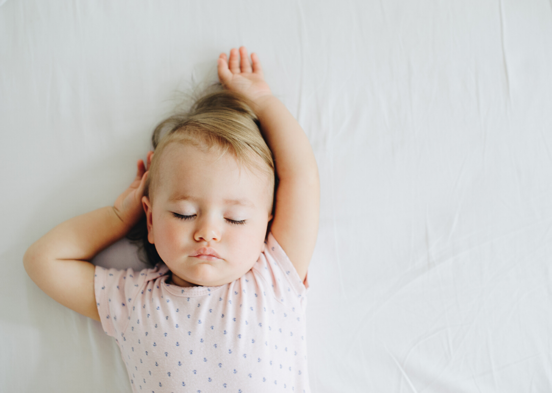 Sleeping baby girl on bed with one arm raised.