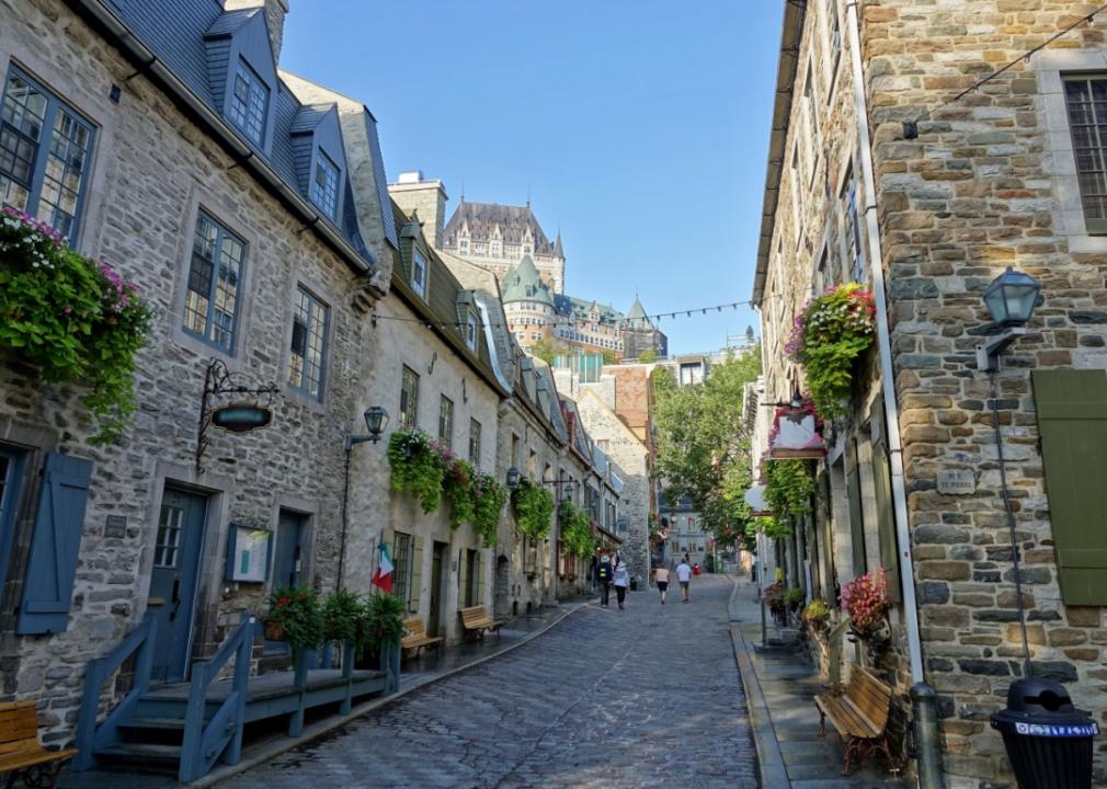 A narrow, historic and cobblestoned street lined with old stone buildings that have a lot of character. Many of the buildings have flower boxes in the windows and hanging baskets overflowing with colorful blooms.