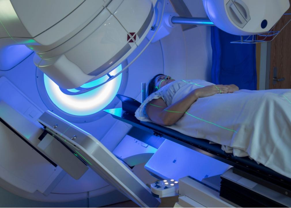 A woman patient covered under white sheets is lying inside a large medical machine emulating blue lights. 