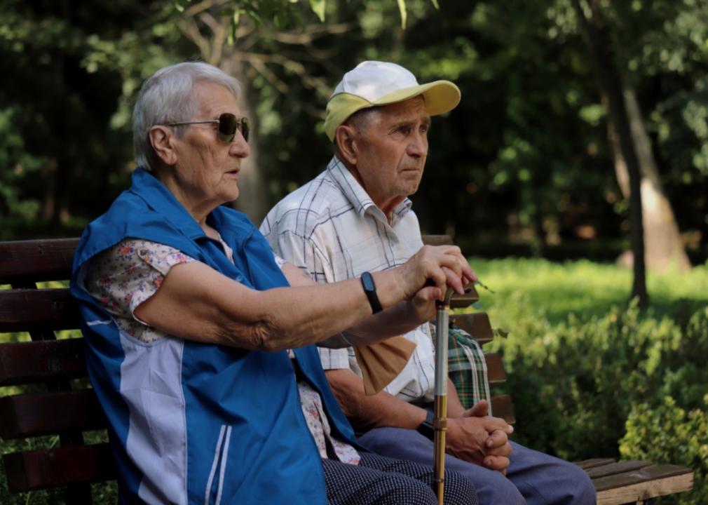 An older couple sitting on a bench in a park. An older woman wearing sunglasses, resting her hands on the cane.