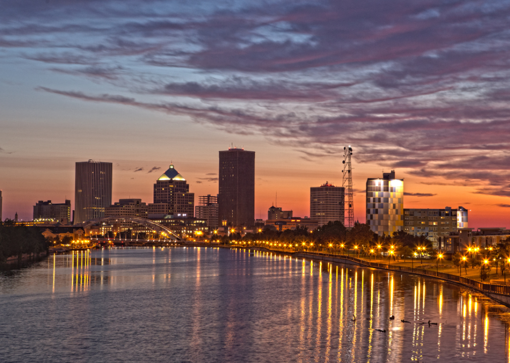 A city skyline with a river in the foreground reflecting vibrant orange hues of sunset sky. 