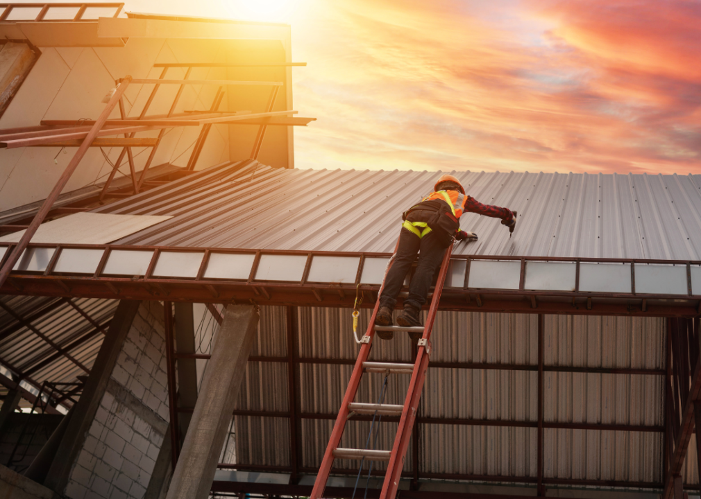 A man standing on top of ladder drilling into the metal sheet on the roof. 