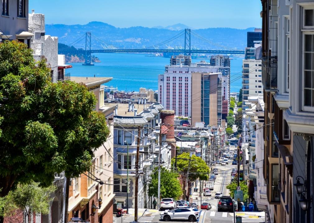 A steep, narrow street lined with colorful houses climbs uphill with cars parked along the road and cable car tracks running through the street and bridge spanning the bay,