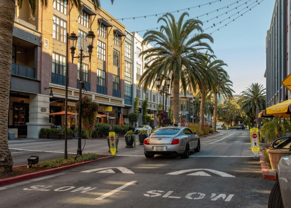 A street lined with palm trees on both sides and elegant buildings featuring clean, modern architecture with large windows and outdoor dining spaces.