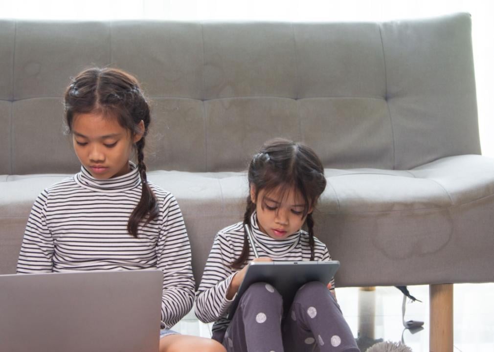 Two Asian girls are sitting on the floor in front of the sofa. The older girl is looking at the laptop and the other one is looking at the iPad.