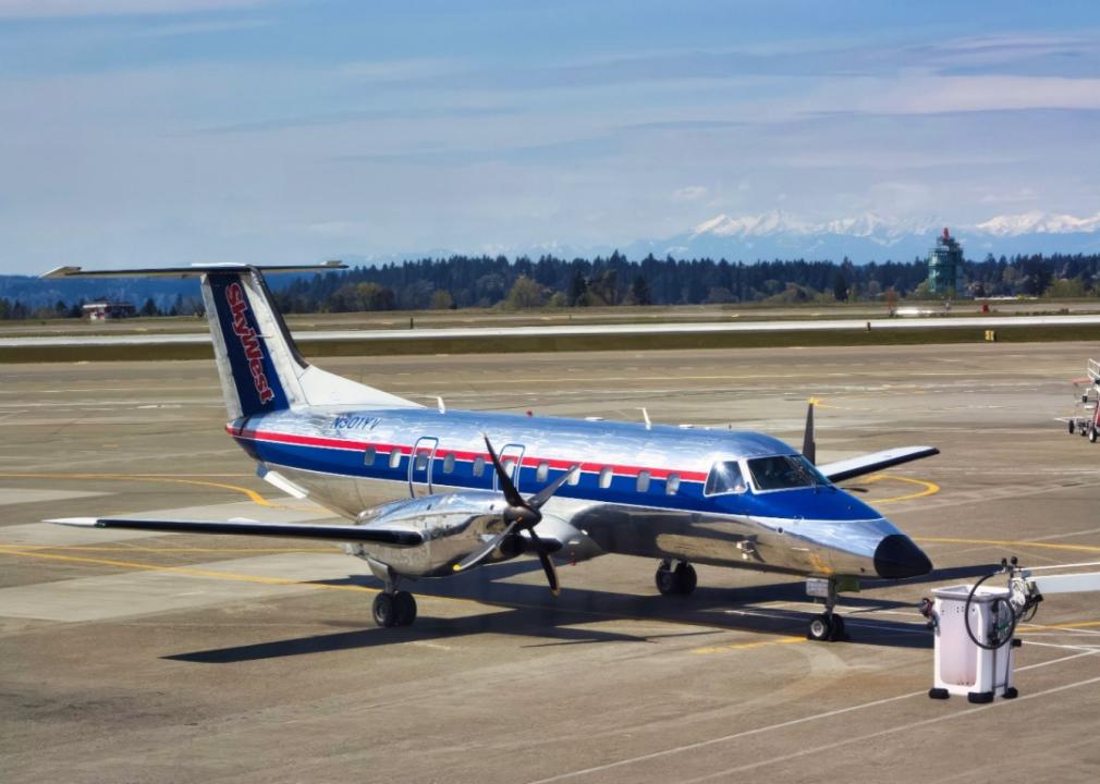 A smller SkyWest Airlines airplane on the tarmac at an airport. The plane is shiny, metallic silver accented by a blue stripe running along the body of the plane with red detailing. The tail is marked with the SkyWest logo.