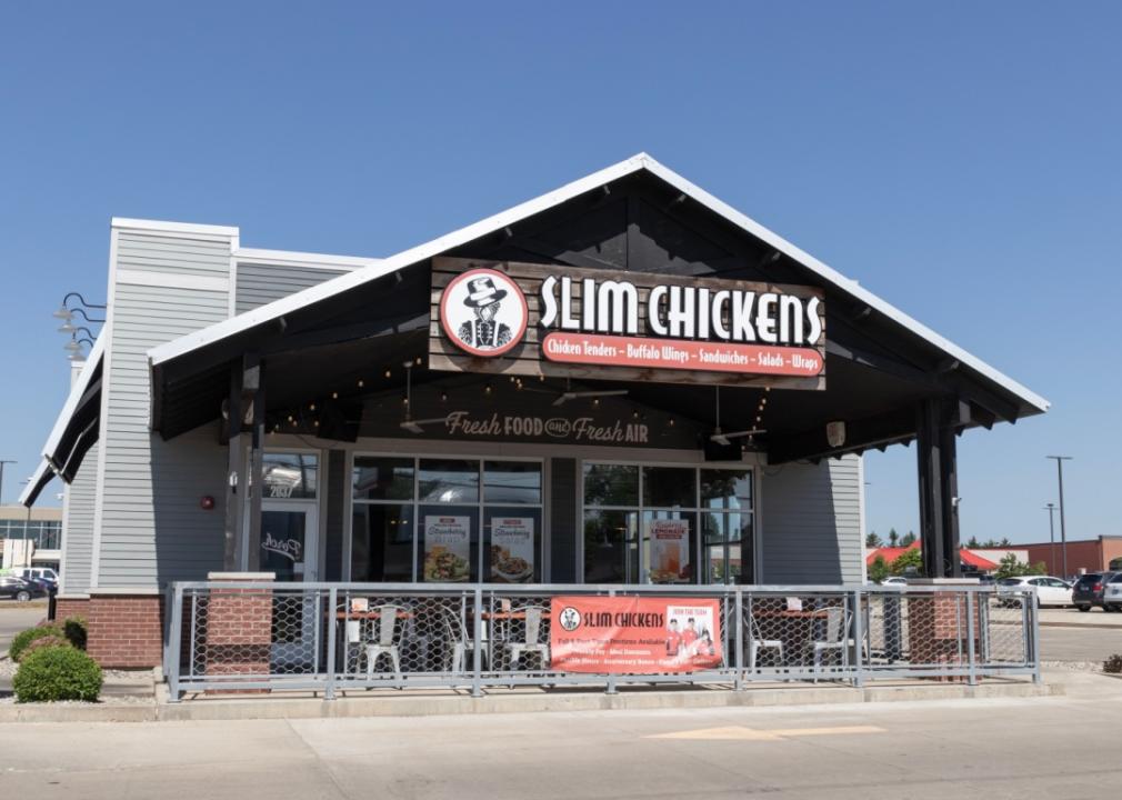 One story, gray building facade with large windows and a large Slim Chickens logo above the entrance. The logo features a chicken holding a red drumstick. There is a fence around the patio in the front with outdoor seating.