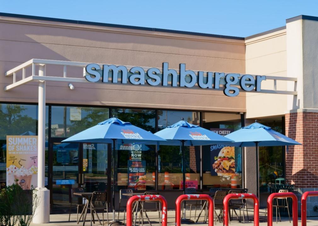 The exterior of a Smashburger restaurant, with several blue umbrella-covered tables out front. 