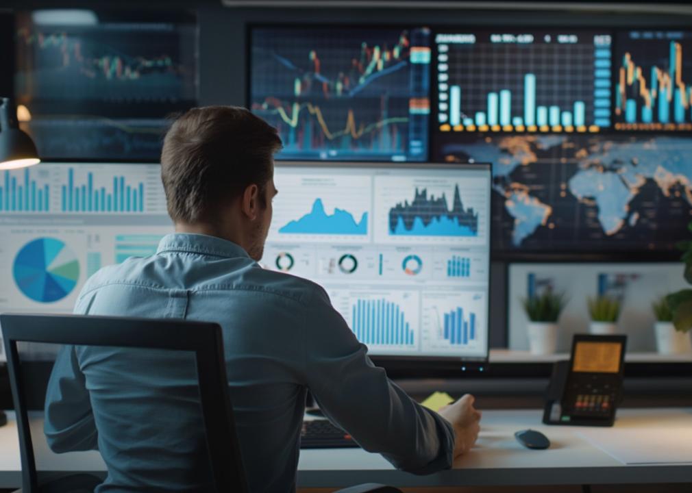 A man seen from behind working at a desk surrounded by multiple large screens displaying various finance- and data-related charts and graphs.