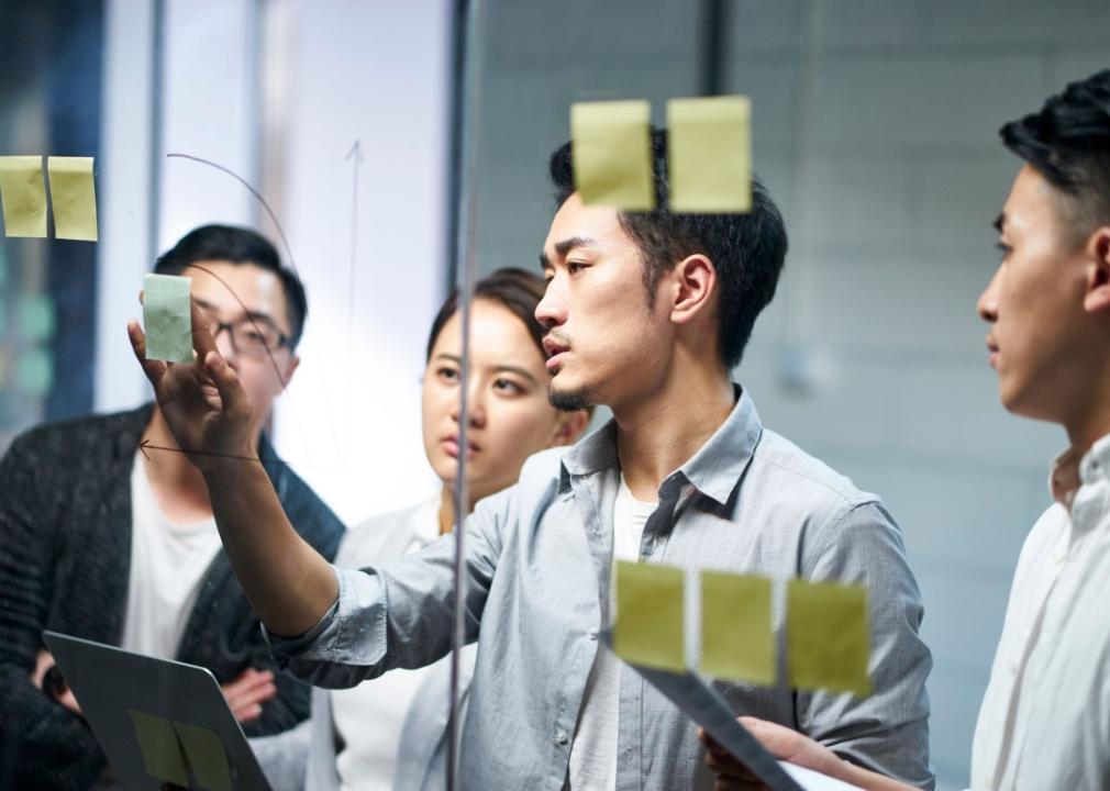 A young man putting an adhesive note on glass while three people also look at the glass board.