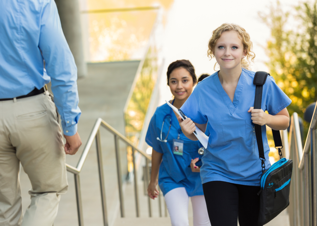 A young blond woman dressed in blue scrubs is walking up the stairs carrying a notepad in one hand and a bag over her left shoulder.  