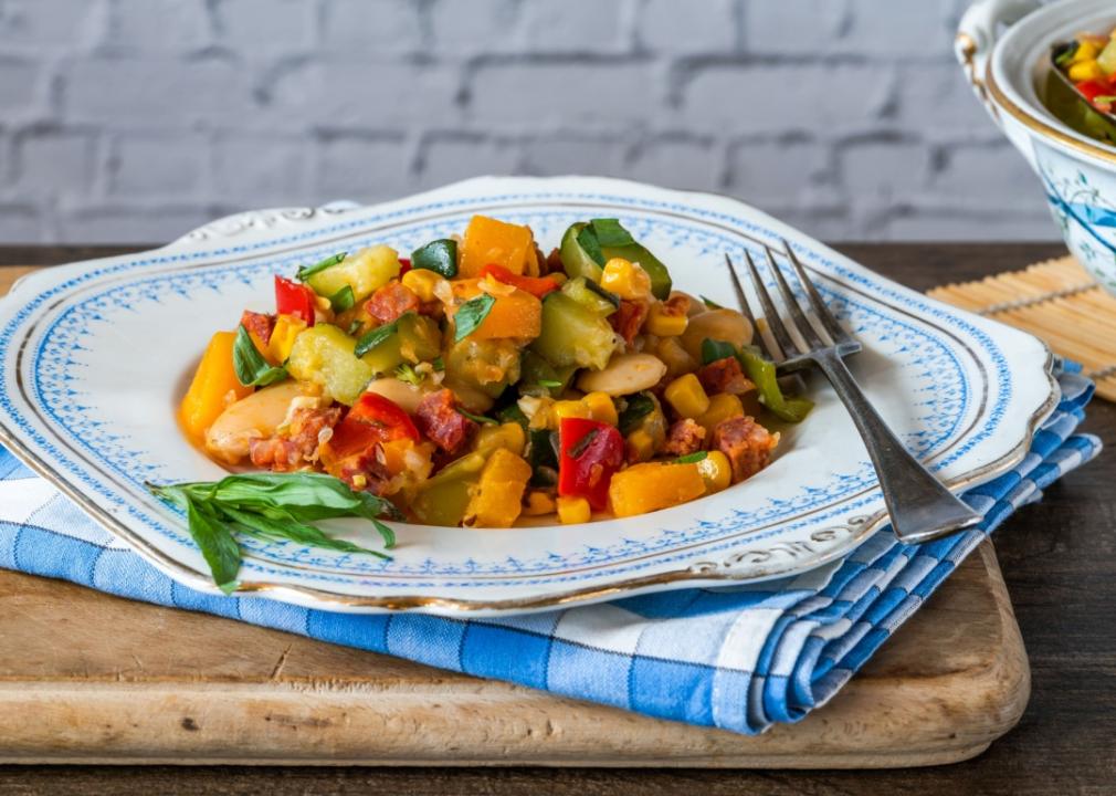 A plate of vegetables, butter beans, butternut squash, sweetcorn and pieces of sausage. The plate sits on top of blue kitchen towel and cutting wooden board.  