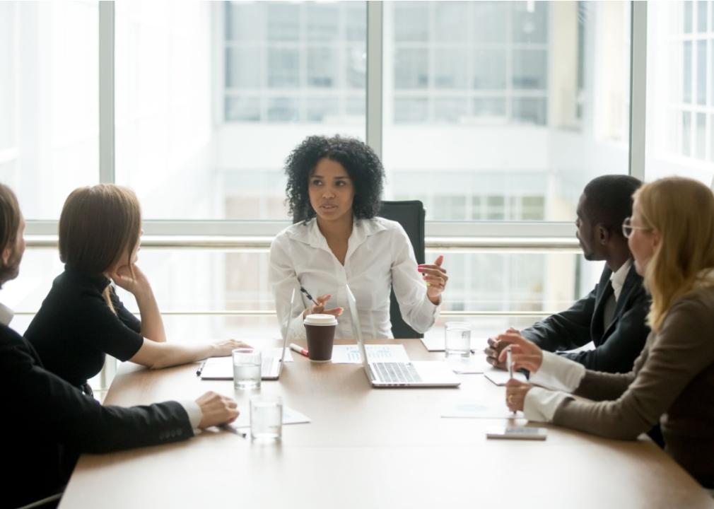 A woman sitting at the front of the table in a conference room looking at a group of people in front of her.