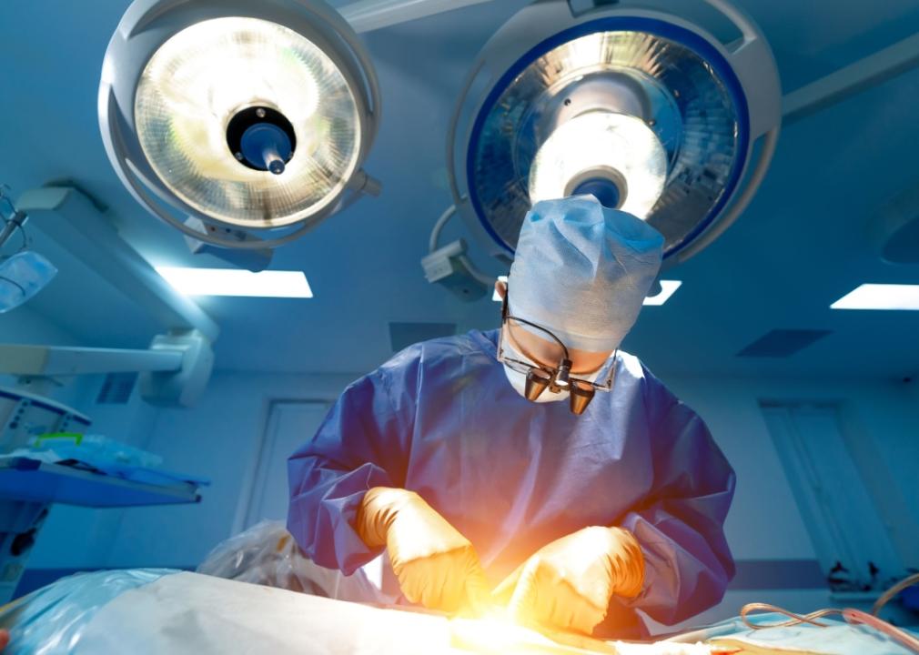 Doctor wearing scrubs and surgical loupe in an operating room looking at the patient lying on the operating table covered under blue drape sheets.
