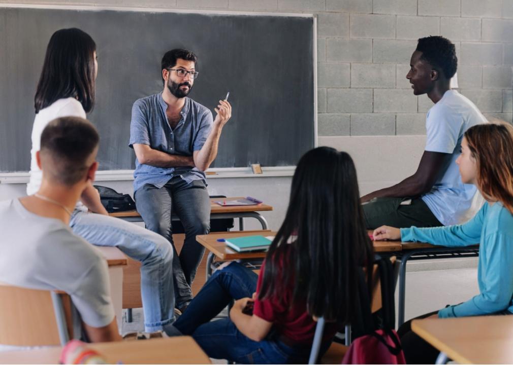 A male teacher sitting on top of the desk looking at the African American male student to his left in a classroom. The rest of the students are facing the teacher. 