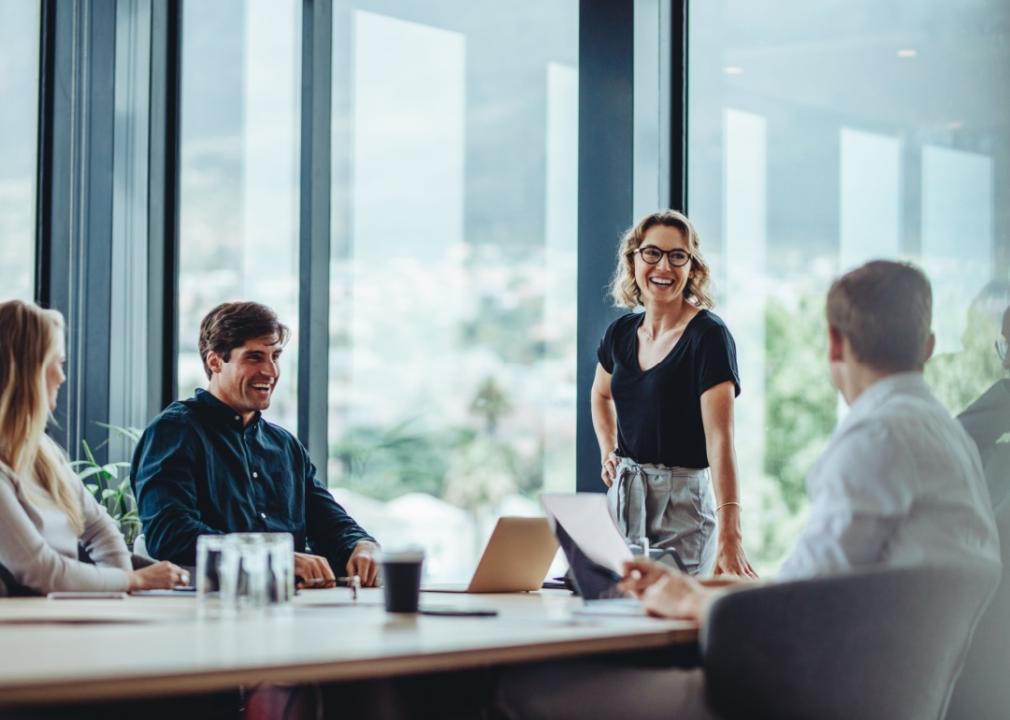 A blond woman smiling standing by the table in a conference room. Three other people are sitting at the table looking at womans direction.