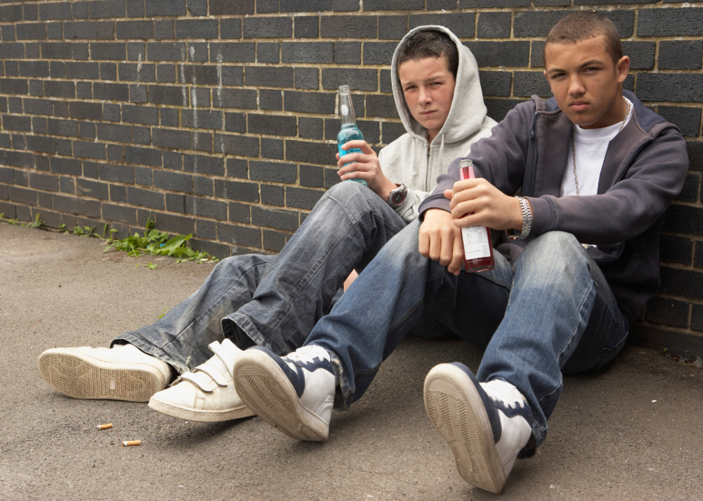 Two teenage boys sitting on the ground against a brick building with bottles of alcohol beverages in their hands. 