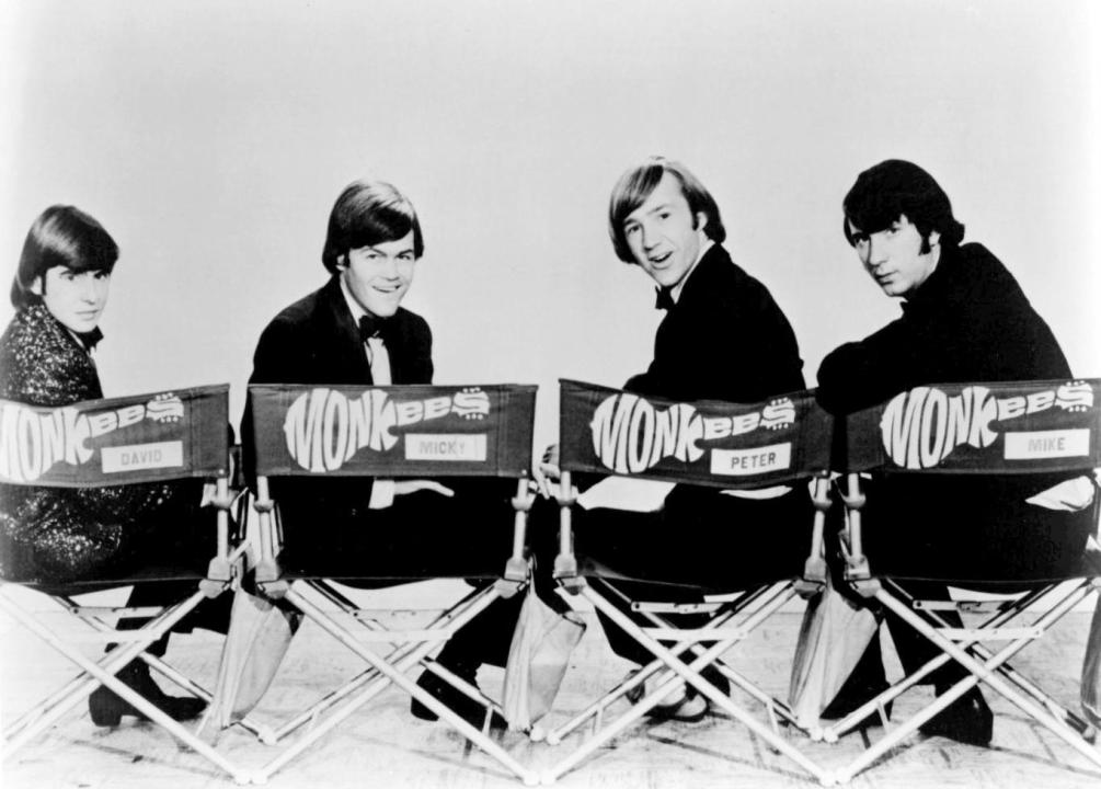The Monkees seated in four director chairs with their logo on the back