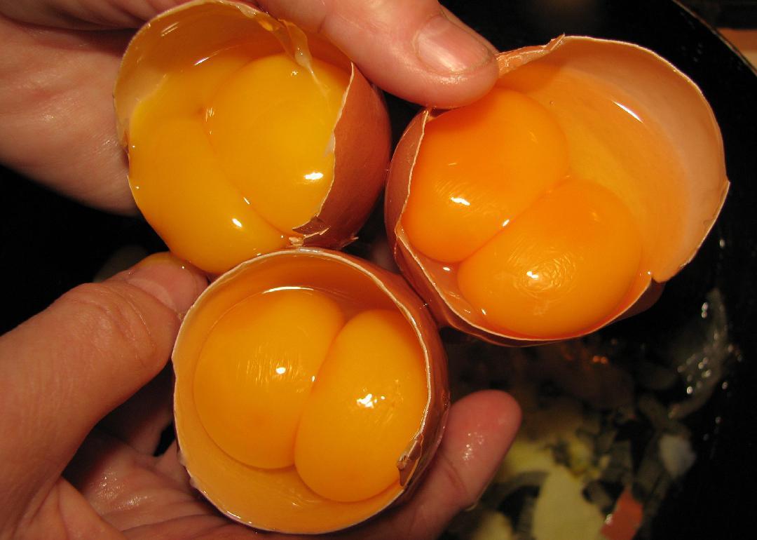 A pair of hands holding three cracked eggs with double yolks. 