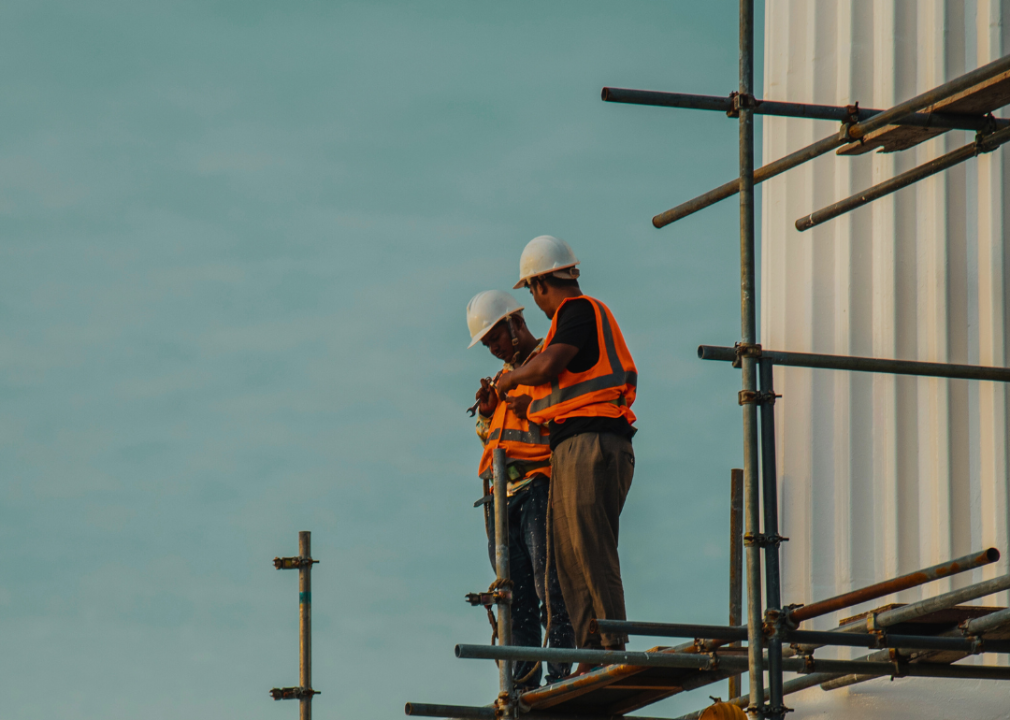 Two men in bright-orange vests wearing hard hats are standing on scaffoldings holding tools.