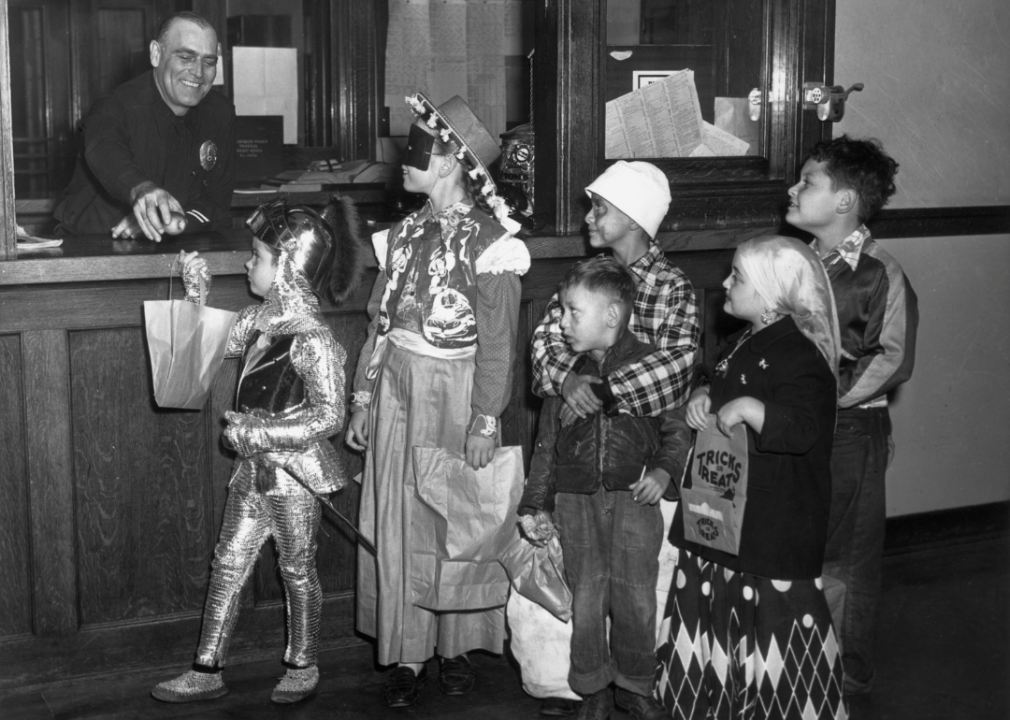 A group of children wearing Halloween costumes wait in a trick-or-treat line to receive apples from an officer at a police precinct.