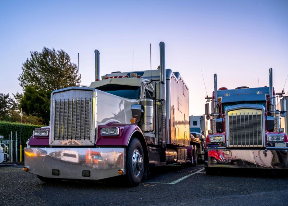 Big rig trucks stand side by side in a parking lot at sunset.