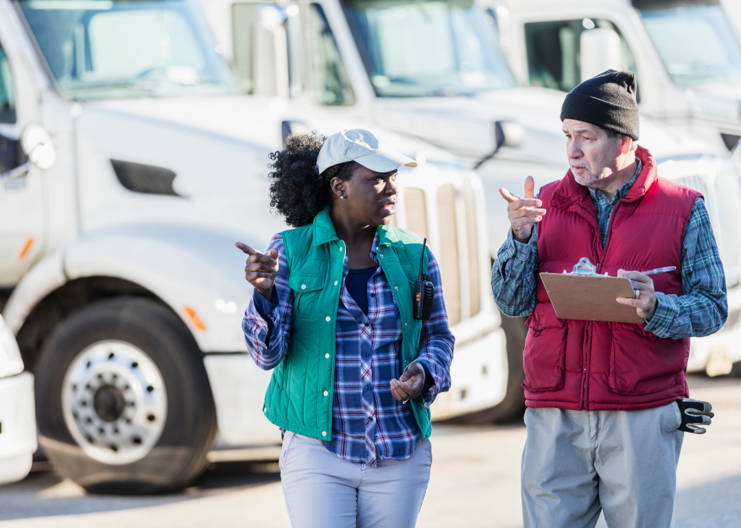 A woman and a man truck drivers in discussion in front a of a fleet of semi-trucks.
