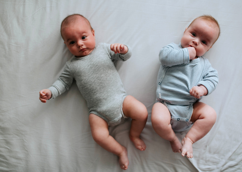 Two baby twin infants are lying down on grey sheet.