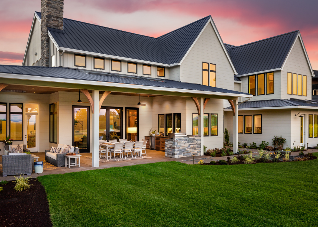 A beautiful, new construction luxury home exterior at sunset, featuring a large covered patio with outdoor furniture, a barbecue, and a table with chairs.
