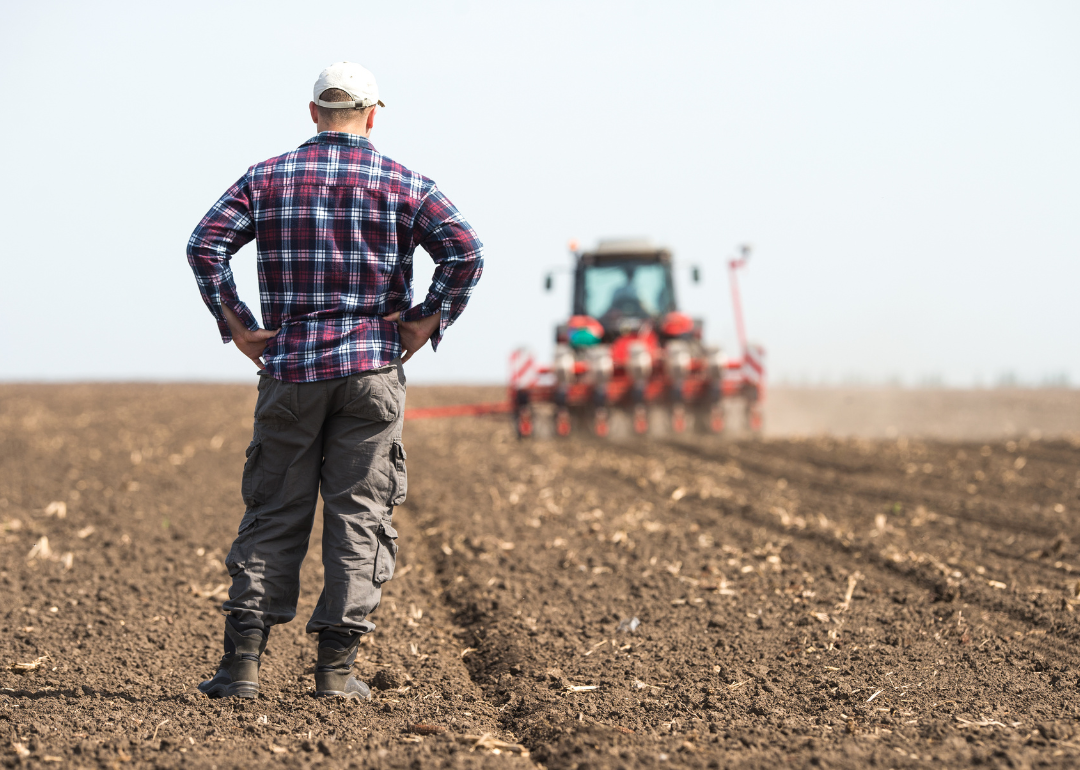 A young farmer standing on a field behind a tractor.