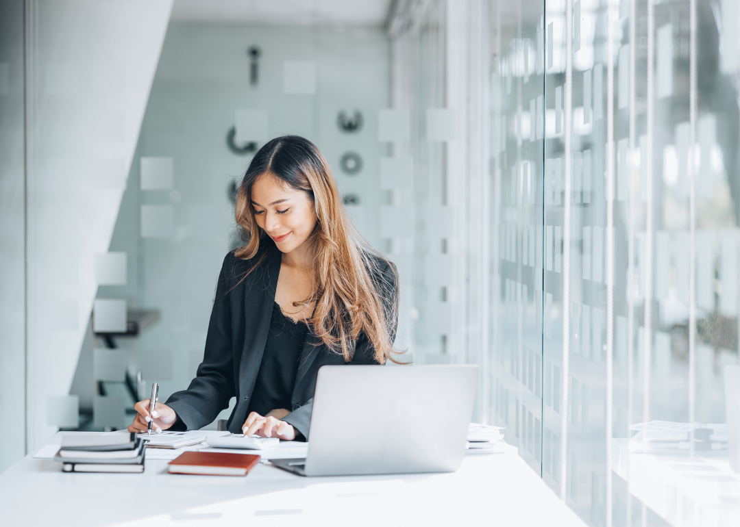 A businessperson sitting in their office writing while on their laptop.