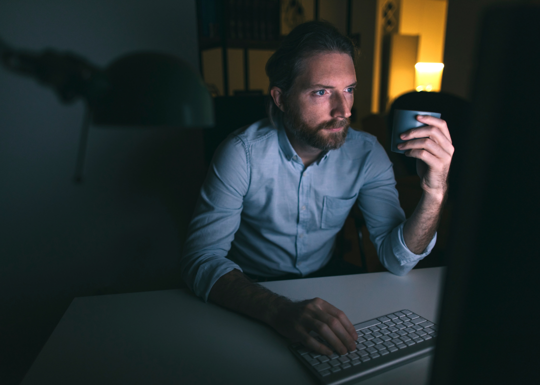 A person working long hours on their computer while holding a mug of coffee.
