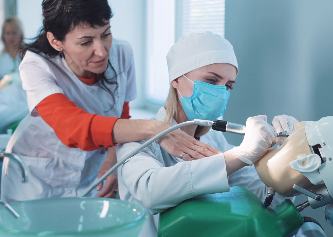 A dentistry student practicing on a dummy at a medical school as a qualified teacher demonstrates the technique to her from behind