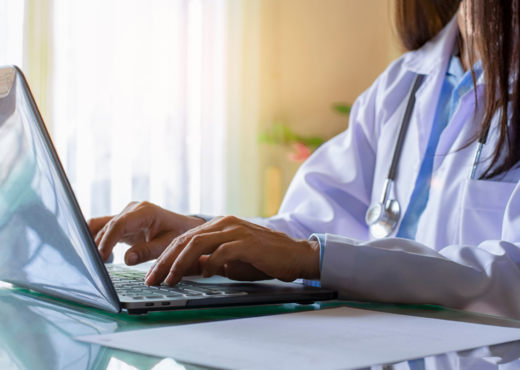 A person wearing a white lab coat and a stethoscope typing on a laptop while sat at a desk. 