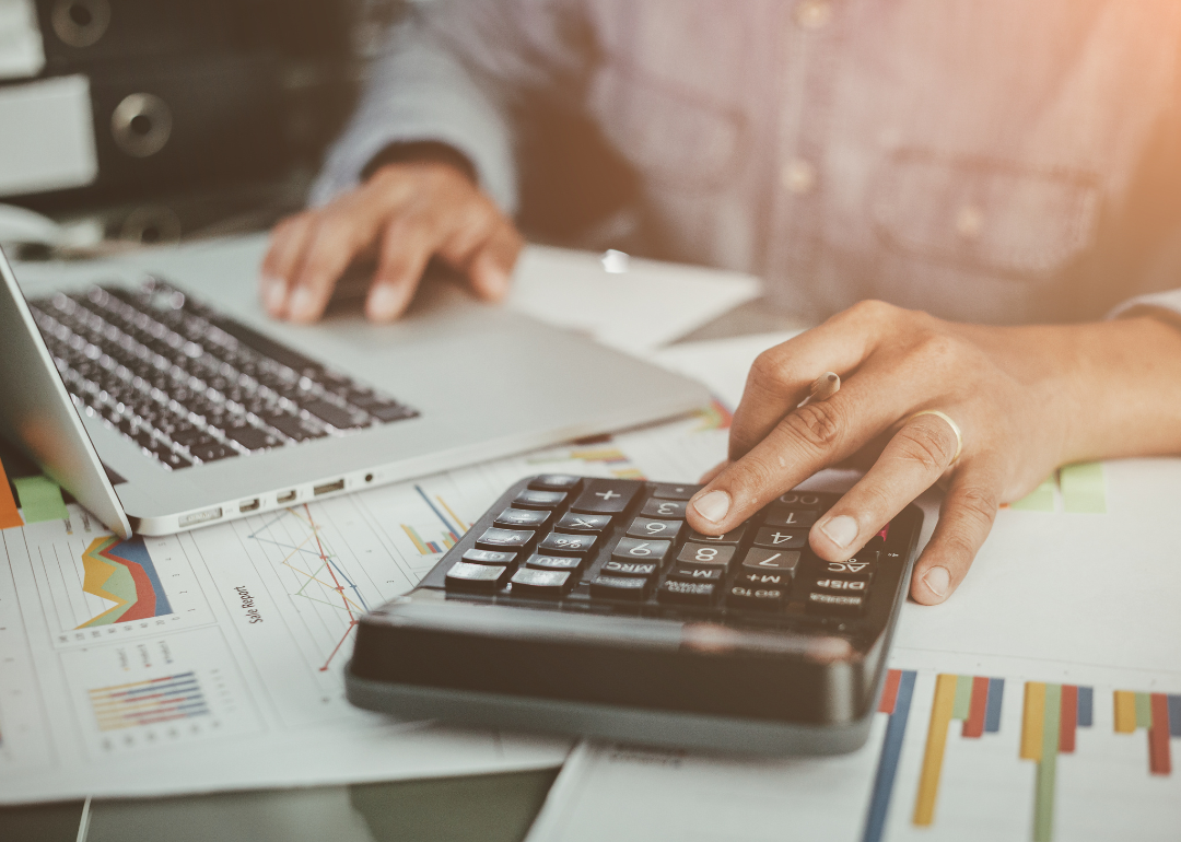 A financial worker scrolling on their laptop with one hand and using a calculator with the other. 