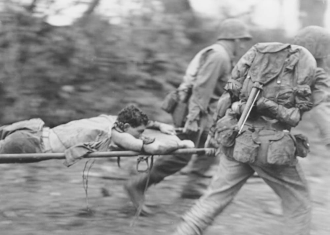 Two soldiers carrying a stretcher where another man is lies prone
