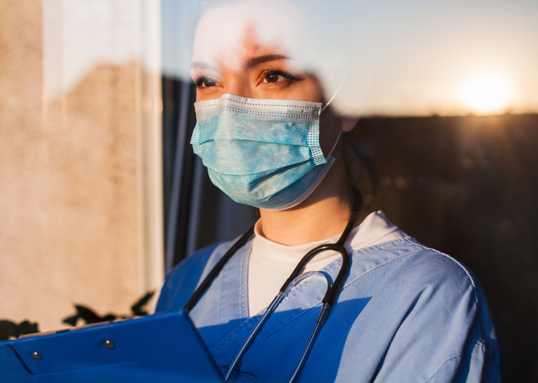 A stressed nurse wearing a mask and looking through a window