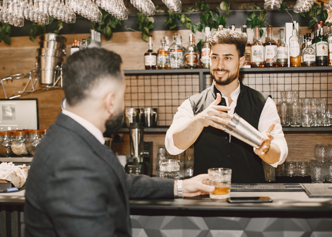 A bartender mixes a drink for a man sitting at the bar.