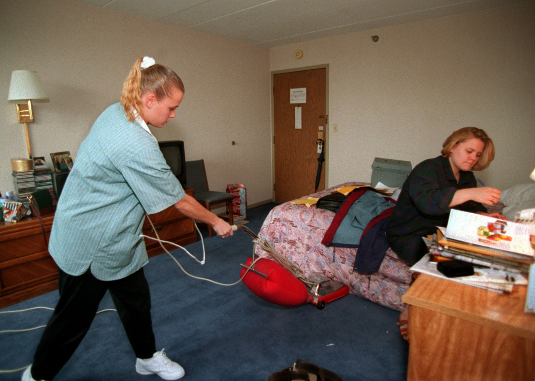 One person working in and one person cleaning a college dorm room.