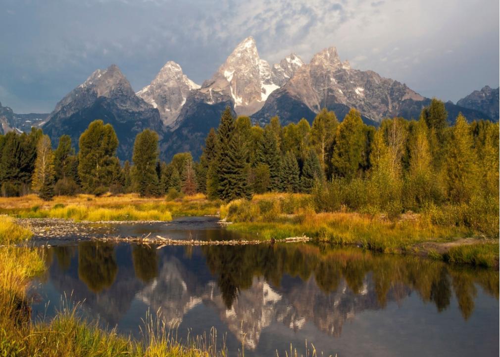Tall, rocky mountain peaks in the background with greeen trees and body of water in the foreground.