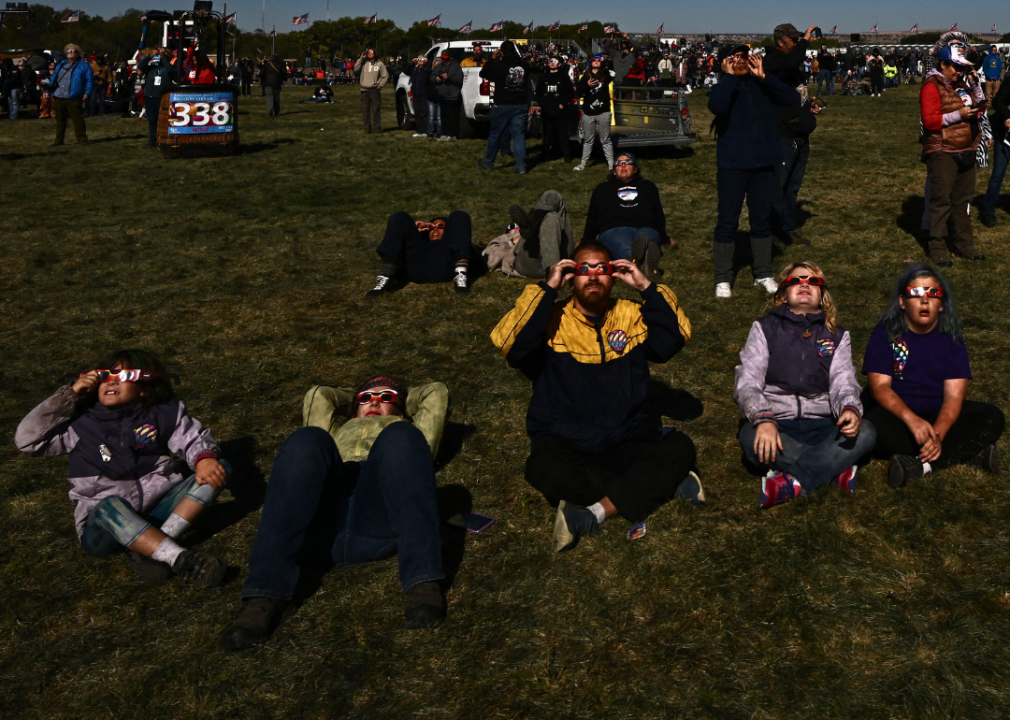 A crowd of people sitting on the grass in large field looking up into the sky wearing cardboard sunglasses. 