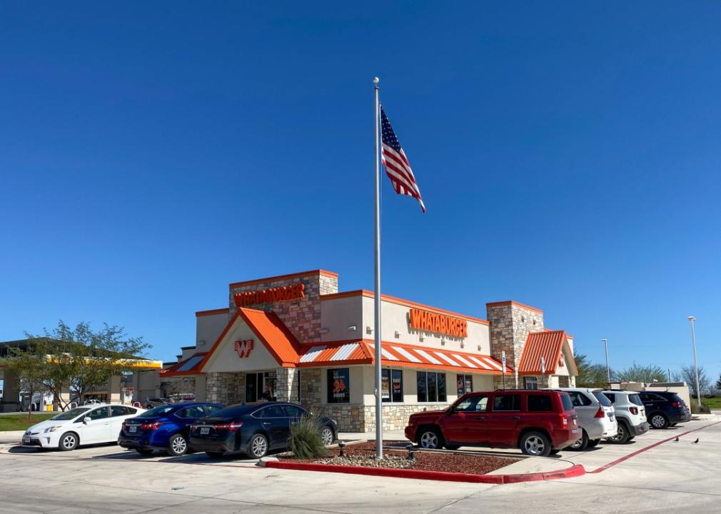 A Whataburger restaurant with a flagpole with an American flag in front of it, and several cars in the parking lot.