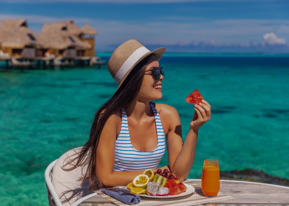 A young woman wearing a straw hat lounges outdoors at a table by the blue waters, delicately grasping a slice of watermelon, with a platter of fresh, vibrant fruit in the foreground.