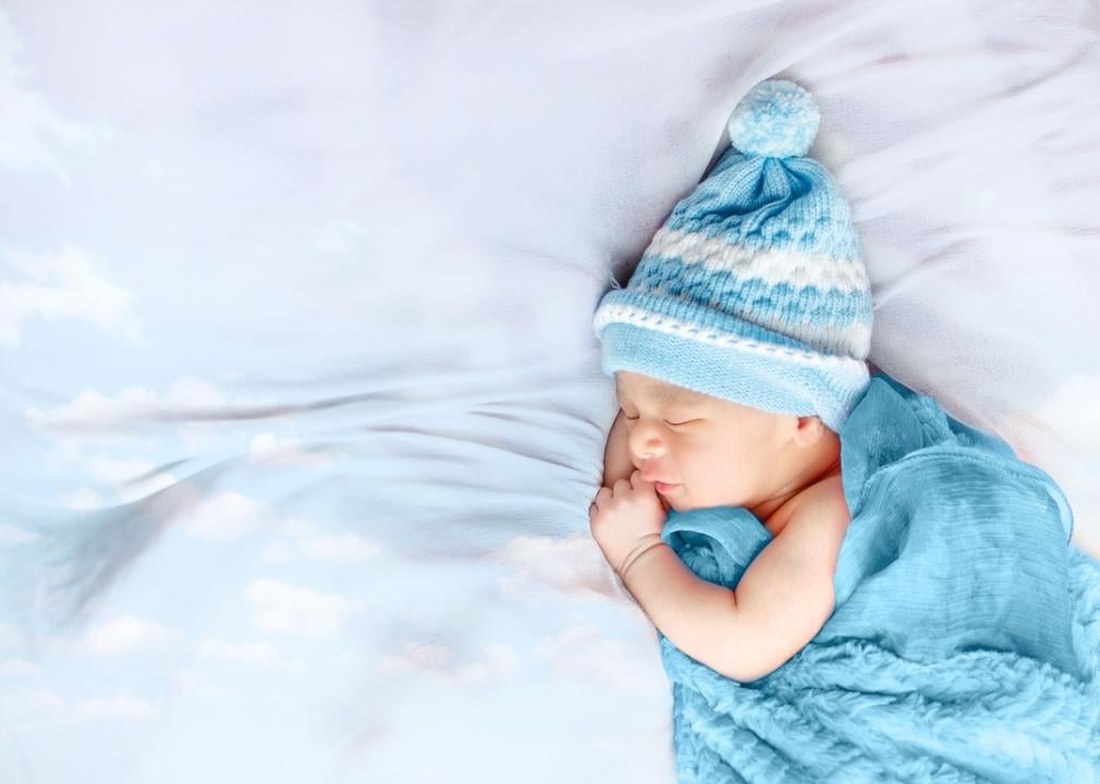 A sleeping baby bundled in a blue beanie with a pom pom and a blue blanket.